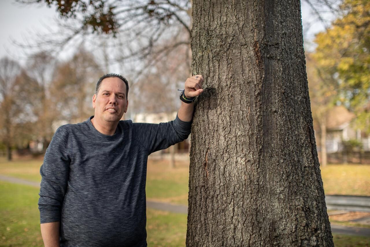 Sean, a Pompe disease patient, smiles at the camera while leaning against a tree